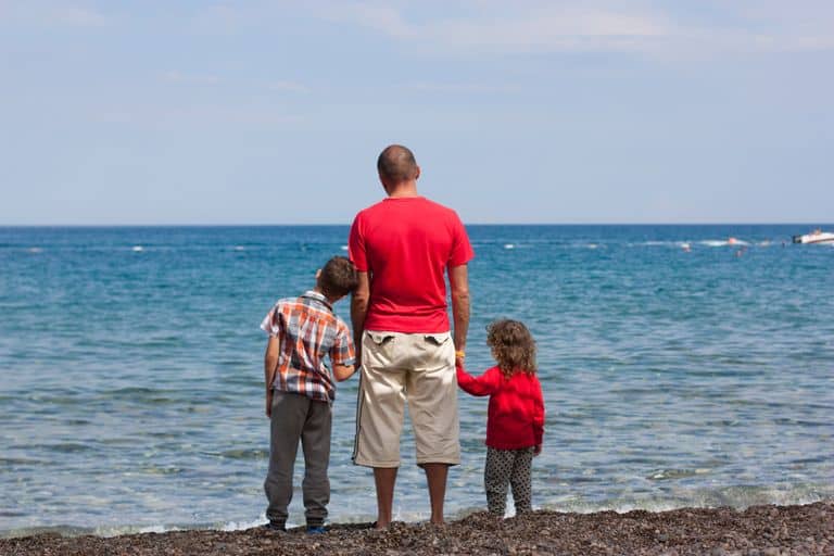 Back view of anonymous young man in casual clothes holding hands of little children while admiring sea during summer holidays